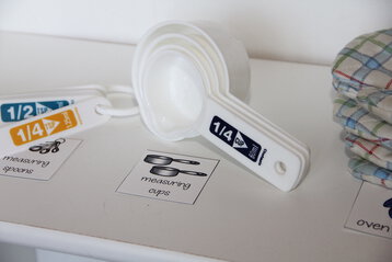 Photo of kitchen supplies (measuring cups and spoons, oven mitts) on a shelf in a dramatic play center. The shelf contains labels to help supplies get put away neatly.