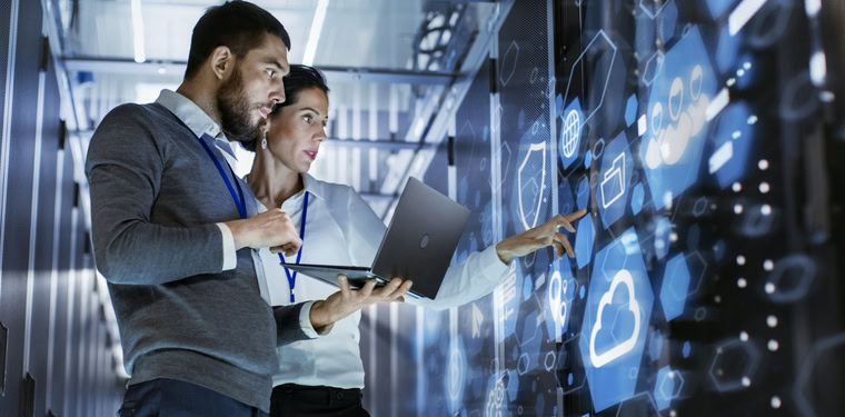 Profissionais em frente a um Rack de rede com os símbolos de uma nuvem, de um escudo e de uma bateria. Representação da prática dos serviços gerenciados de TI
