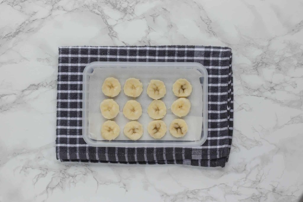 Banana slices lined on parchment paper in a Tupperware container