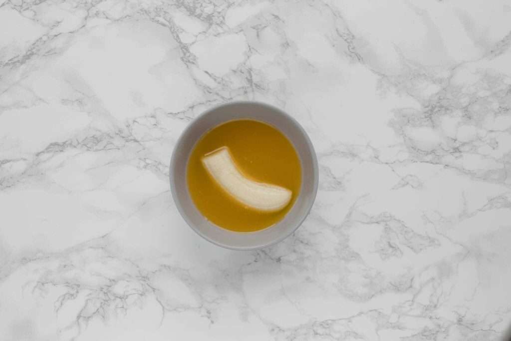 A peeled cut banana submerged in a bowl of orange juice