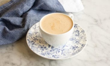 Butter Chai Latte in a white teacup sits on a blue and white saucer on a marble counter. A blue tea towel sits in the background.