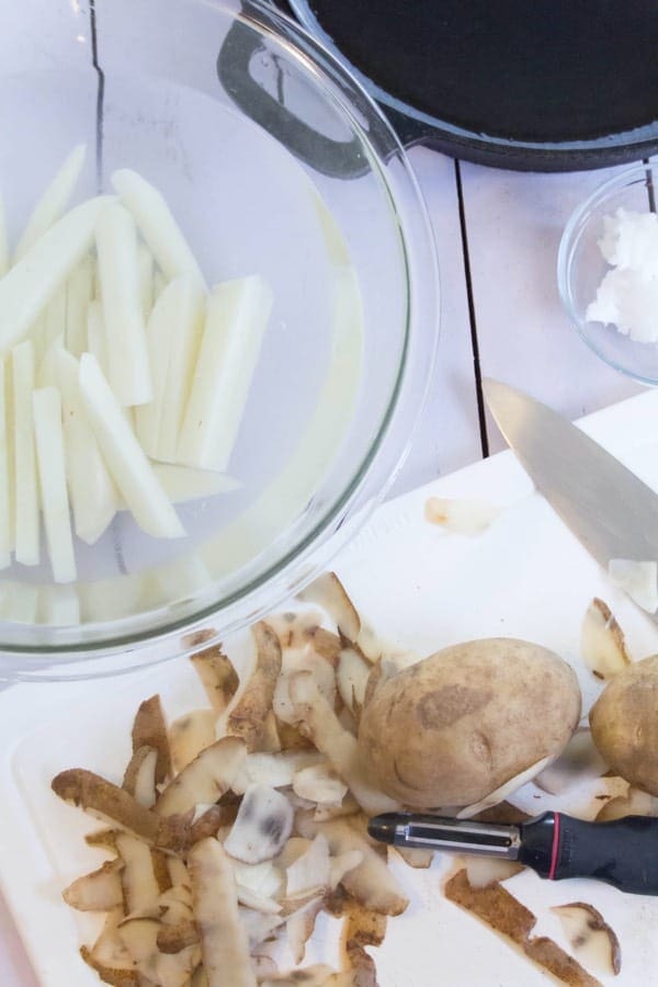 raw potatoes on cutting board with some cut potatoes in a bowl, with a knife and frying pan in the shot.