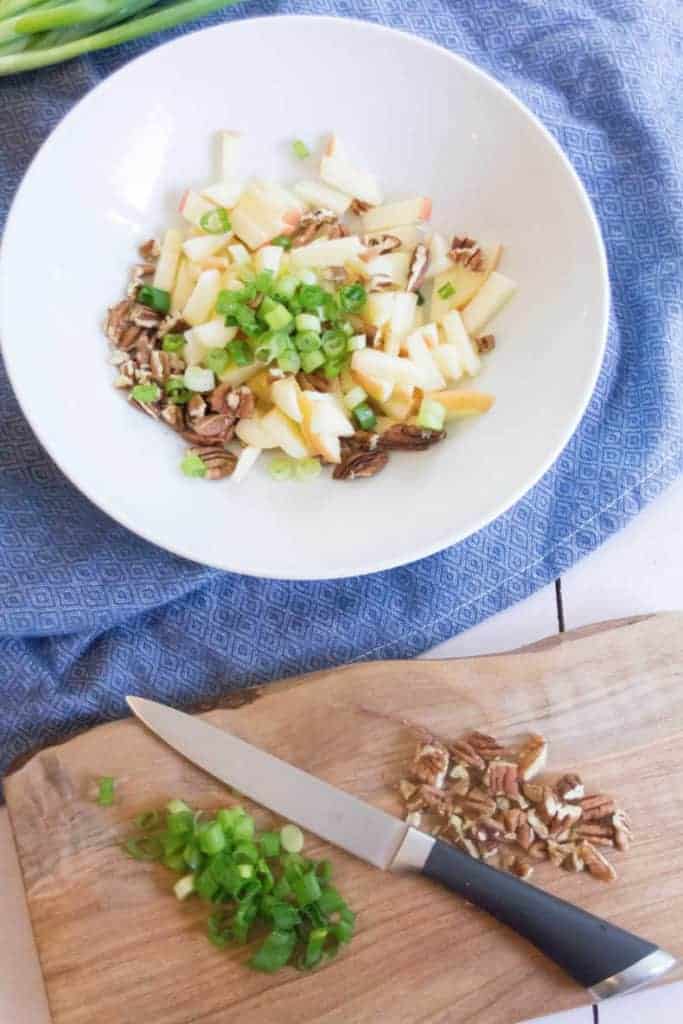 Apple, green onions, nuts, and parsley are tossed together in a medium-sized bowl on a blue tea towel. A wooden cutting board with chopped nuts and green onion sits by the bowl.