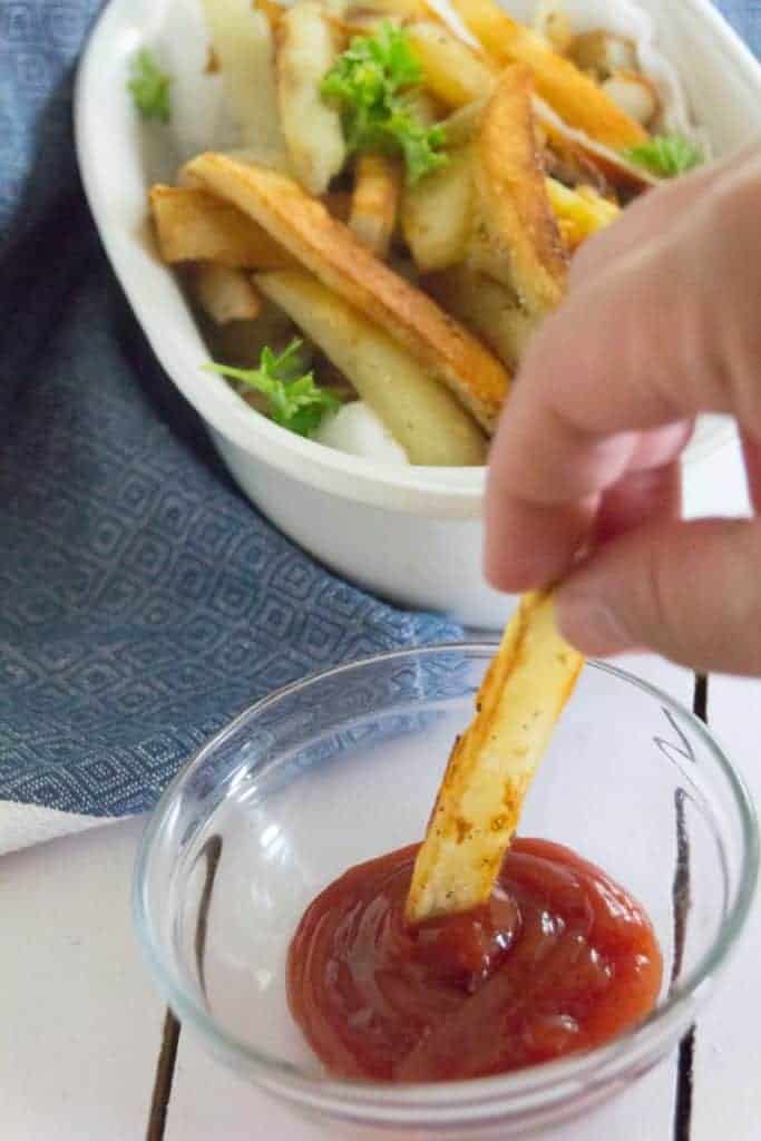 Homemade french fries in a white bowl with a hand holding one and dipping into ketchup.