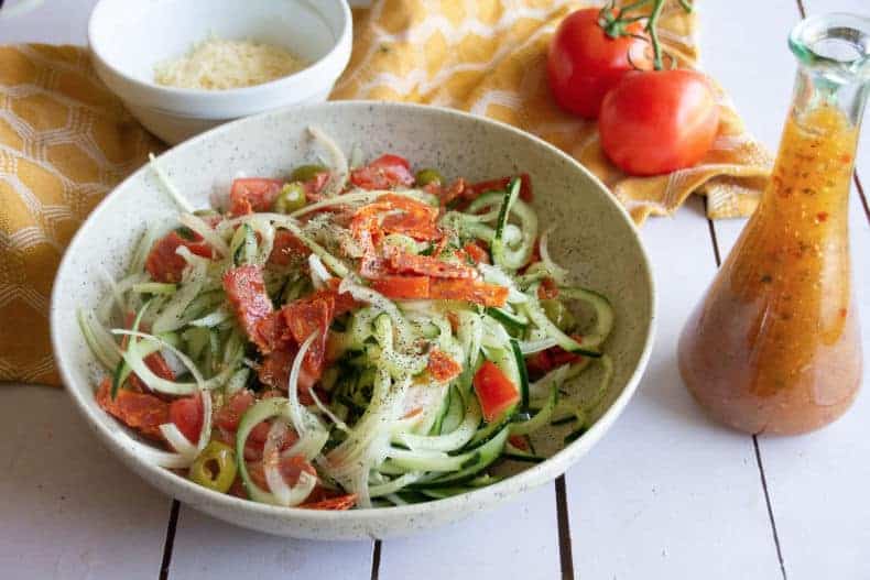 Image of zesty Italian cucumber salad in a speckled bowl on a wood slat table top. Bottle of Italian dressing is beside the bowl, a yellow tea towel, Parmesan and whole tomatoes are in the background