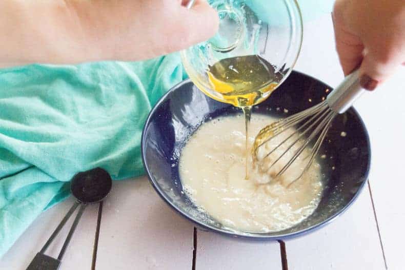 pizza dough ingredients added to a bowl. A measuring cup is pouring oil into the yeast mixture while a whisk incorporates it.