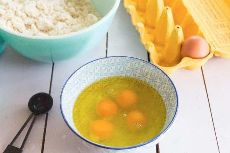 Four eggs broken in a light blue bowl. An empty egg carton and bowl of dough ingredients in the background.