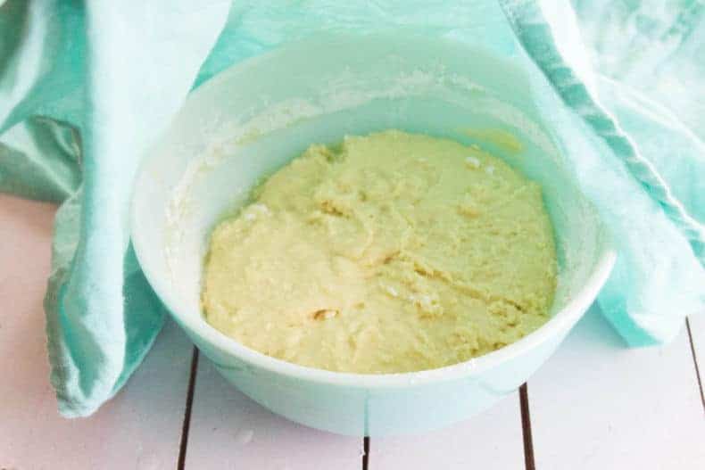 A mint coloured tea towel is lifted up to see the Pizza dough rising in a mint coloured bowl