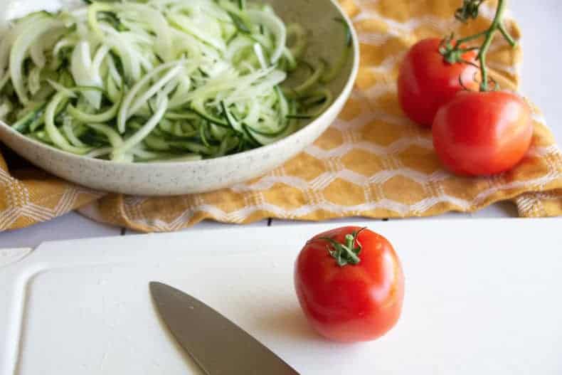 A whole tomatoe sits on a white cutting board with a knife laid next to it. In the background is a white pottery bowl with spiralized cucumbers in it.