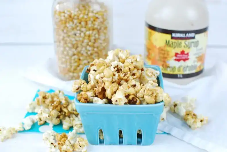 a square ceramic teal basket with homemade stovetop kettle corn spilling out, with a mason jar of popcorn kernels and a jug of real maple syrup in the background