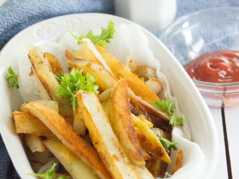 Homemade French Fries sit in a white ceramic bowl, on a blue tea towel on top of a white wood table top. A glass bowl of ketchup sits next to it. A white salt shaker, shakes salt above it.