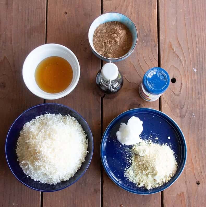 Ingredients for chocolate coconut macaroons laid out on a wooden table: coconut flakes, coconut oil, almond meal, vanilla, honey, cocoa, and salt.