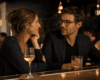 Two adults sit at a bar, leaning toward each other and sharing a quiet, attentive moment over drinks at night.