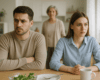 Man and woman sit at a kitchen table with serious expressions, an older woman stands behind them in a doorway.