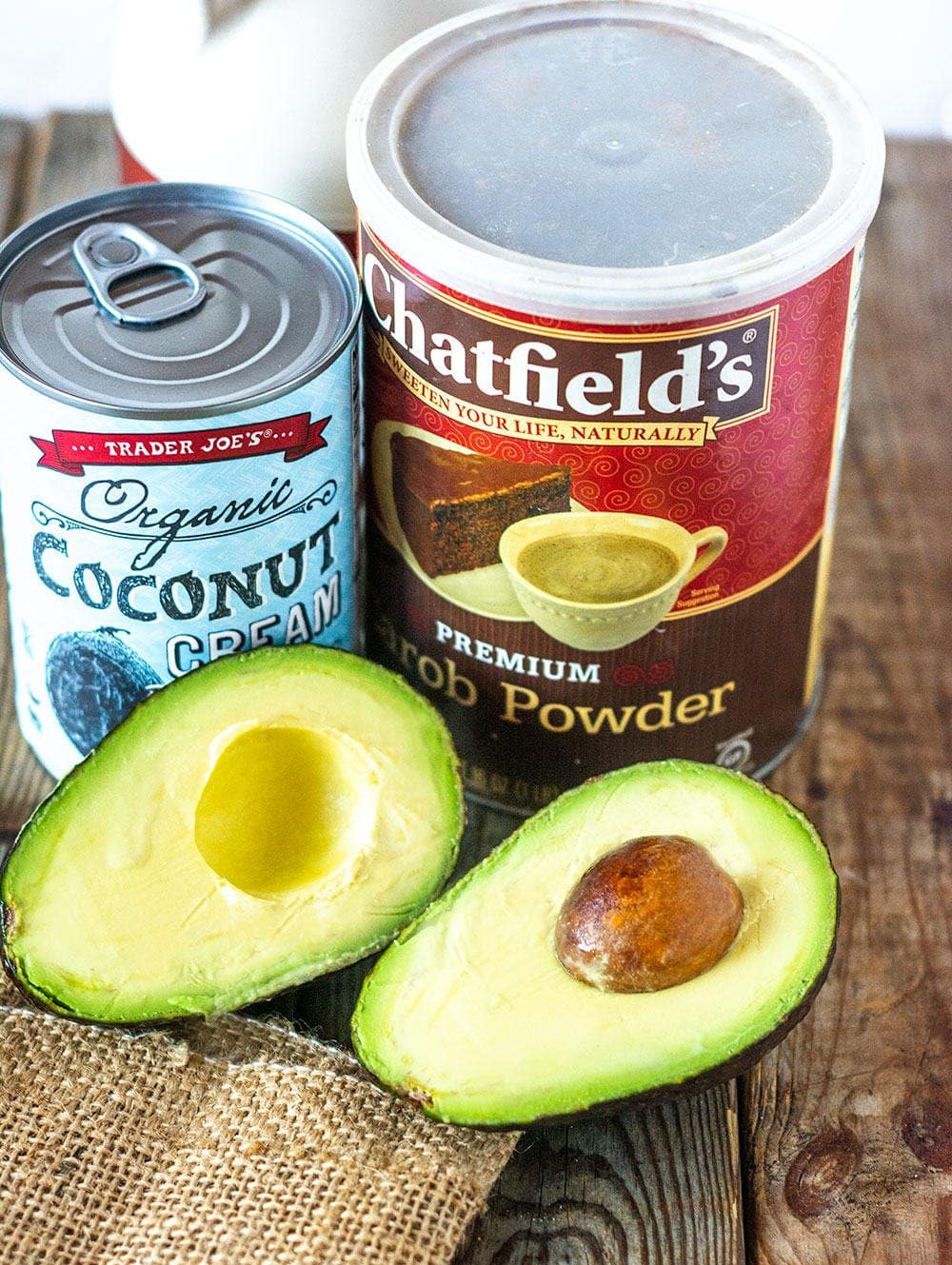 Carob Mousse ingredients, jar of carob, avocado, coconut cream can, maple syrup jar on a wooden background