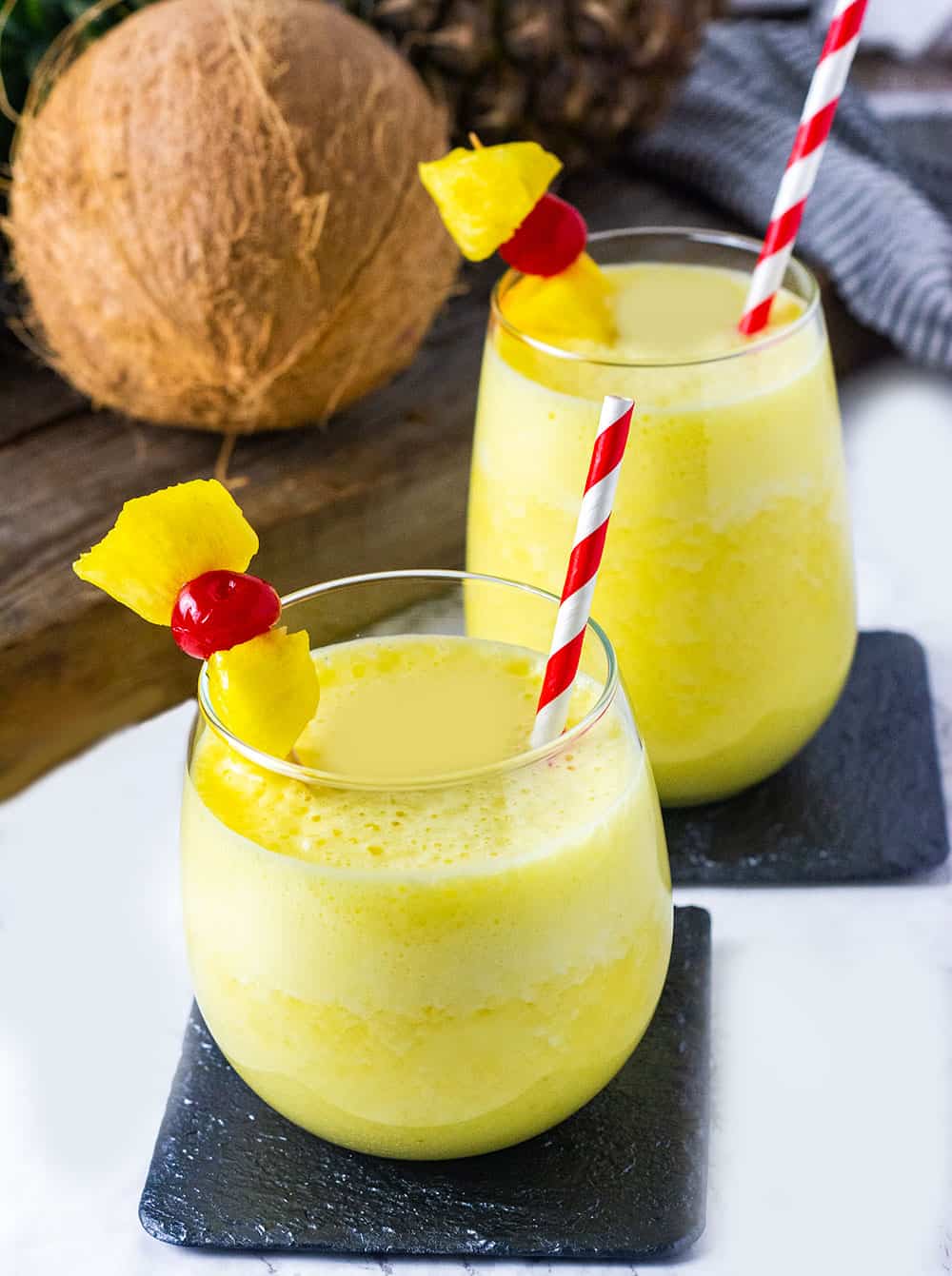 Easy Pina Colada Smoothie, 2 glasses of smoothie with pineapple and cherry garnishes, coconut and pineapple in the background