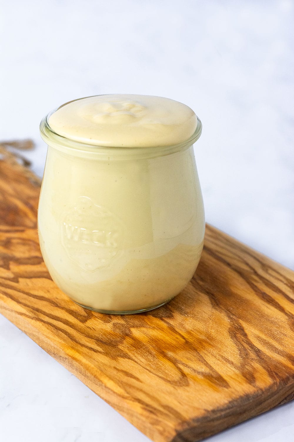 Vegan mayo recipe in two glass jars the one in the foreground with a silver spoon, the one in the background on a cutting board