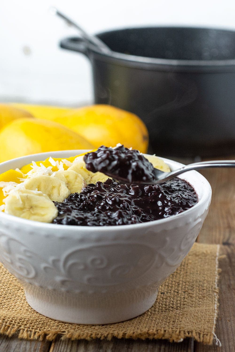 forbidden rice pudding in a white bowl, one spoonful being taken, garnished with banana and mango slices with mango in the background