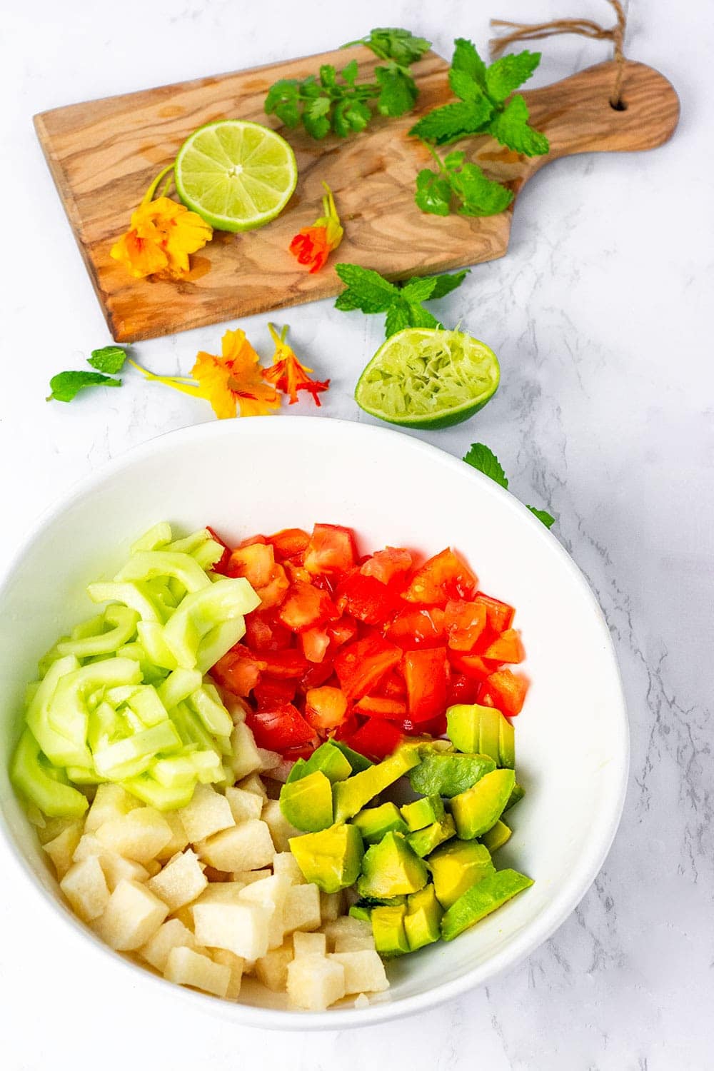 Jicama salad overlay with ingredients in a bowl, cubed jicama, tomato, cucumber and avocado with cutting board in the background with herbs