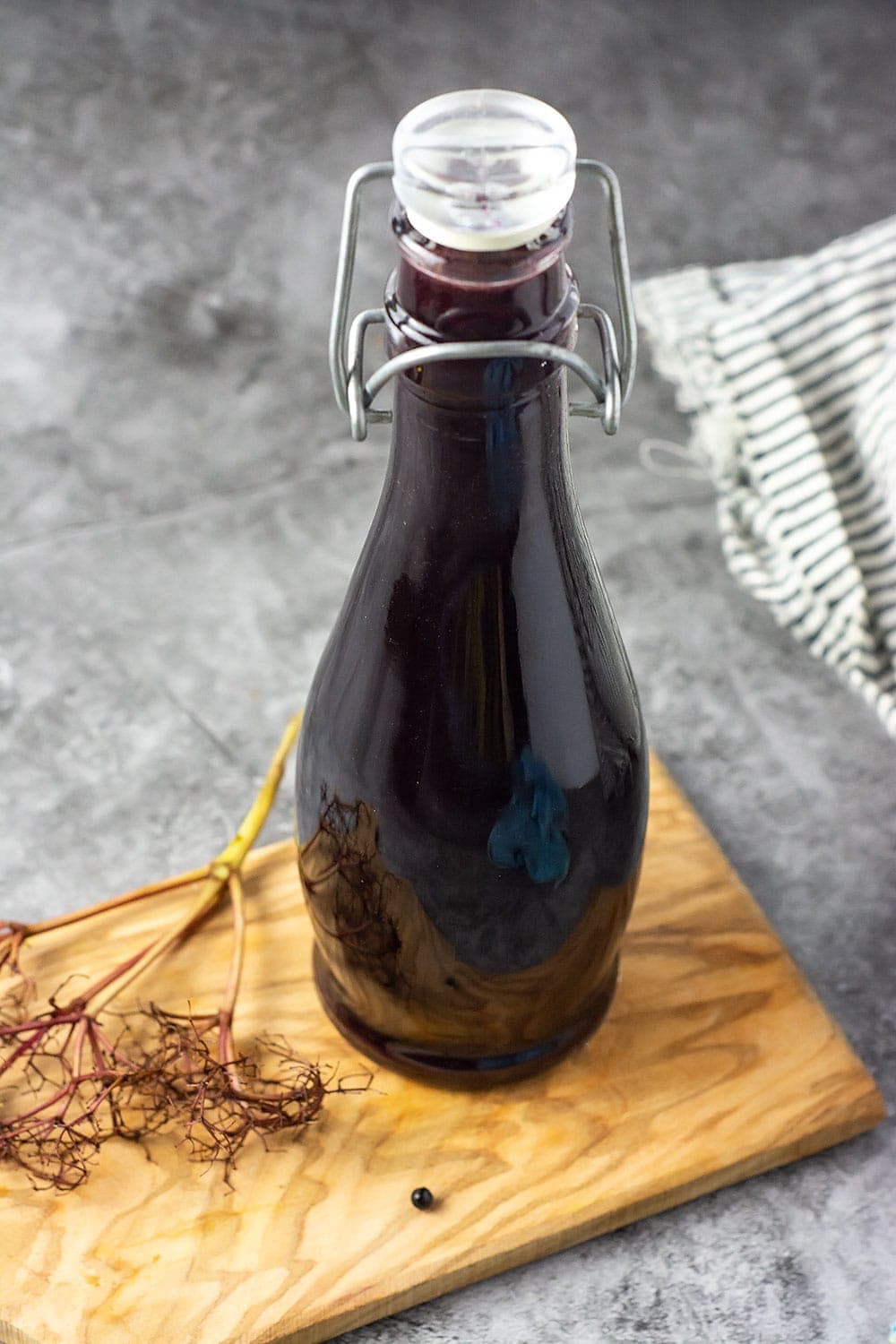 Elderberry syrup recipe in a glass jar on a wooden cutting board with a stem of elderberry on a concrete background