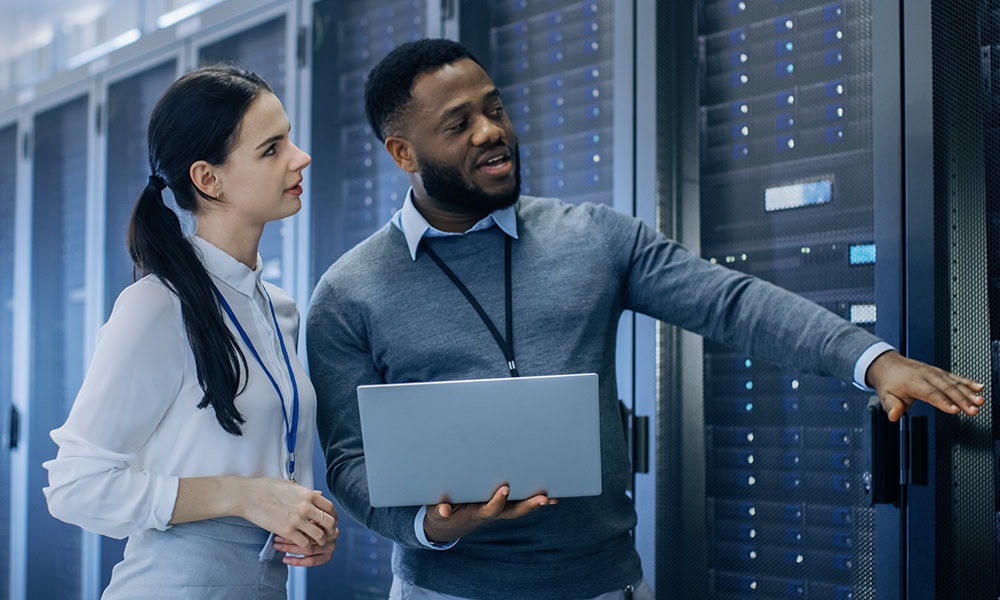 IT technician with a laptop computer gives a young intern a tour. They chat in the data center as they walk next to the server racks. They are performing maintenance work.