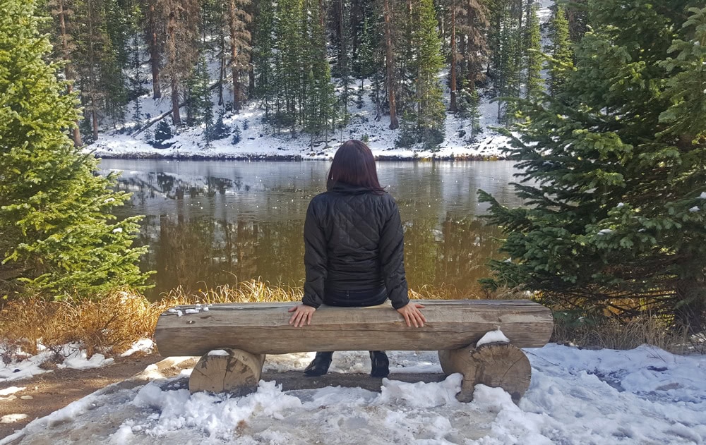 Brooke sitting at a bench looking at the lake by a snow covered trail in Rocky Mountain National Park