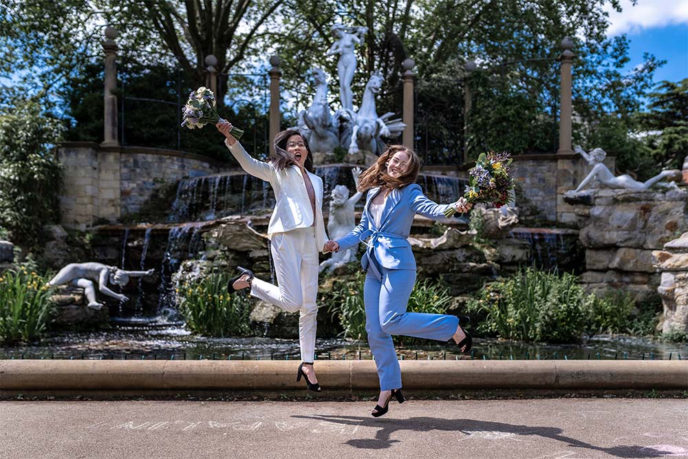 two brides same sex couple jumping with job with bouquets in front of a fountain