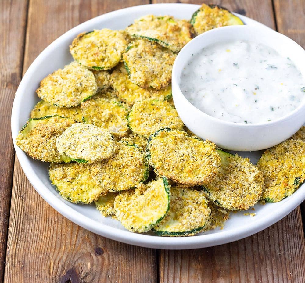 Air Fryer Zucchini Chips on a white plate topped with a small white bowl with vegan ranch dip on a brown wooden board