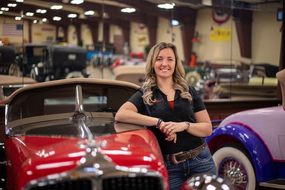 Woman in a black WyoTech shirt leaning again a read car.
