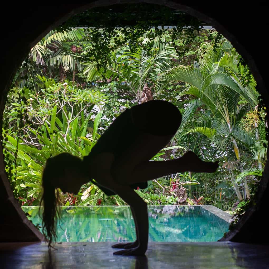 girl practicing crow yoga pose in front of pool and jungle background in Bali