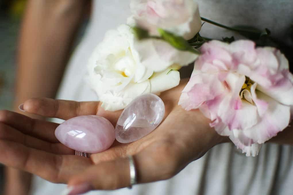 Female hands holding two yoni eggs made from pink quartz and transparent violet amethyst with white flowers