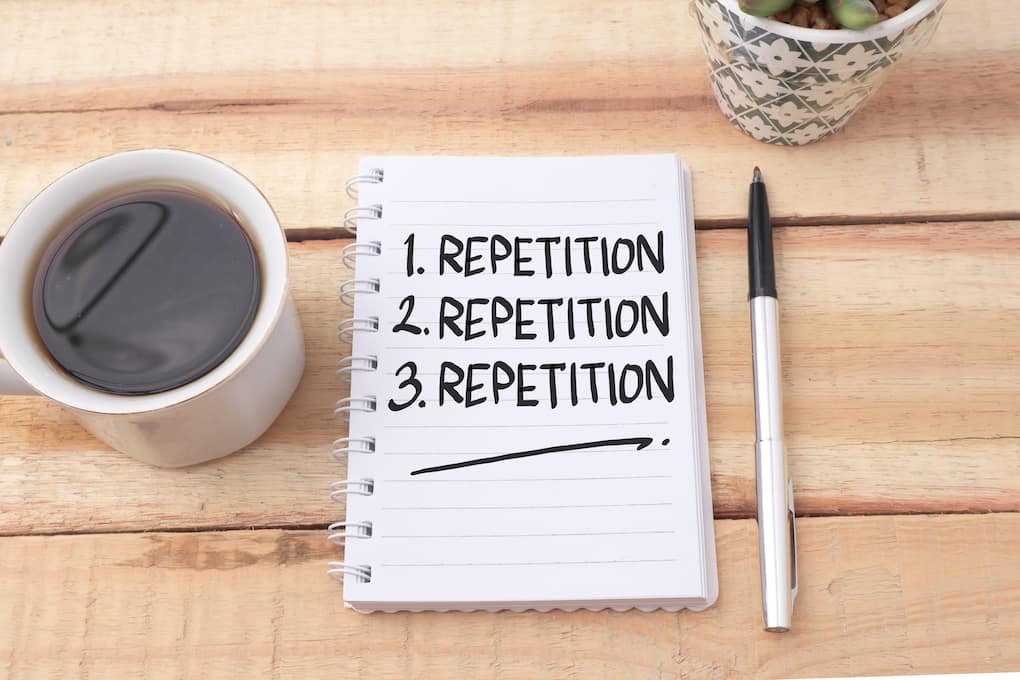 the word repetition written three times on a notepad on a wooden table next to a cup of coffee and a pen