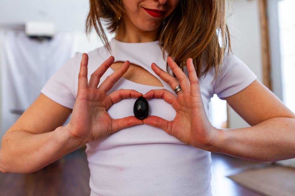 woman holds black obsidian yoni egg to her chest in a heart shape with her hands