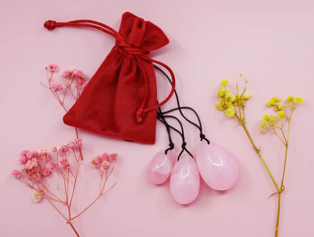 three rose quartz drilled and stringed yoni eggs with red pouch surrounded by flowers