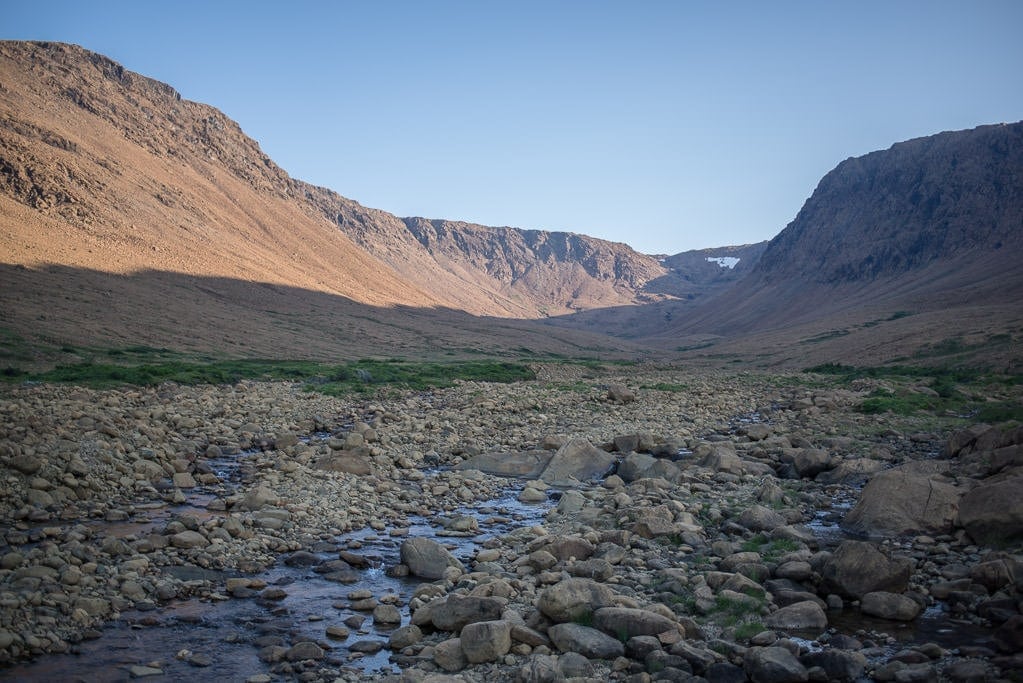 Hiking along the water towards the waterfall with some snow visible on the mountain tops in the distance