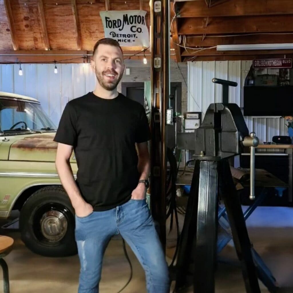 Man in brown beard, black t-shirt, and blue jeans, smiling at the camera. He is standing in a garage with a green, rusty truck behind him. To his left is a black vice. Behind everything is a work bench against the wall of the garage.