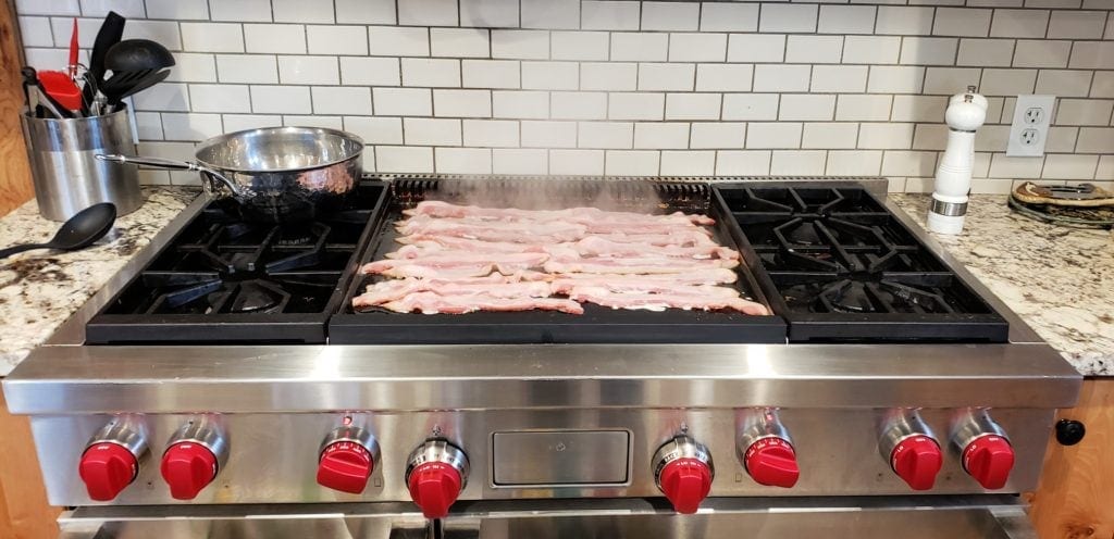 bacon sizzles on the huge flat frying grill on Wolf stainless steel stove with red knobs