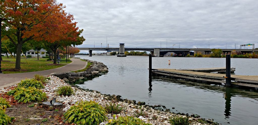 Paved trail meanders between orange maple trees and the riverbank. A dock and bridge frame the watery image of Fox River Trail, one of best Things to do in Green Bay Wisconsin