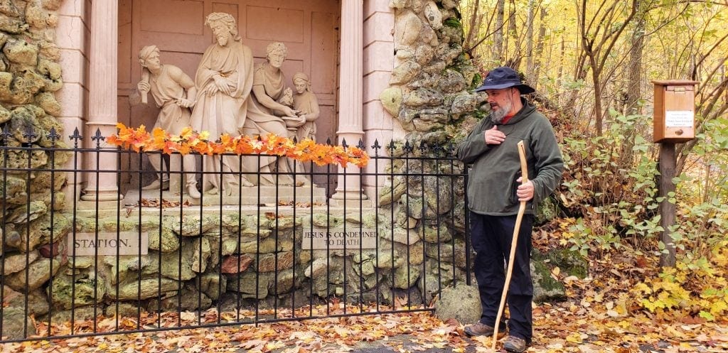 Man with black hat and wooden walking stick taps his heart in front of one of the Stations of the Cross at Holy Hill Church Wisconsin