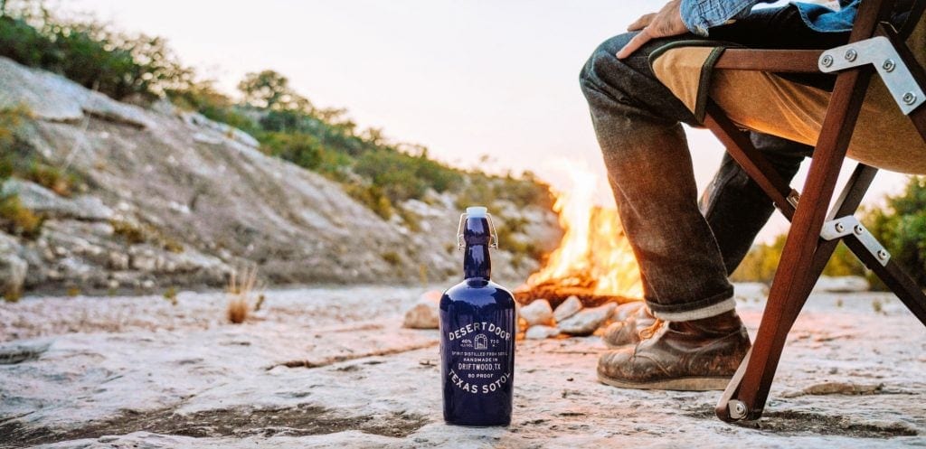 blue bottle of sotol sits on desert floor with a campfire and an sitting in camp chair behind the bottle of Texas sotol
