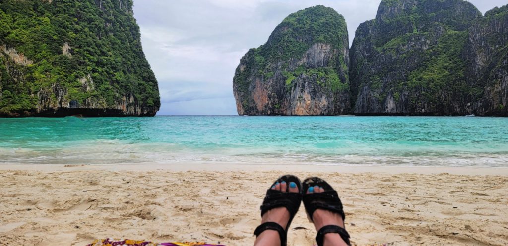 feet with blue pedicure, in black sandlas rest on white sand beach with view of turquoise waters and floating islands of green