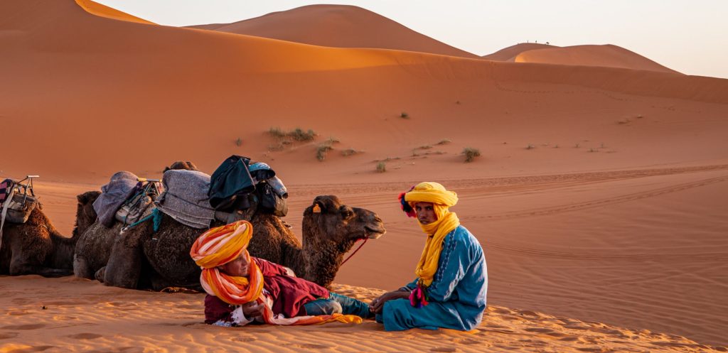 two people in yellow and orange turbans sit in sand with 3 camels laying down-dunes in the background in Morocco tips