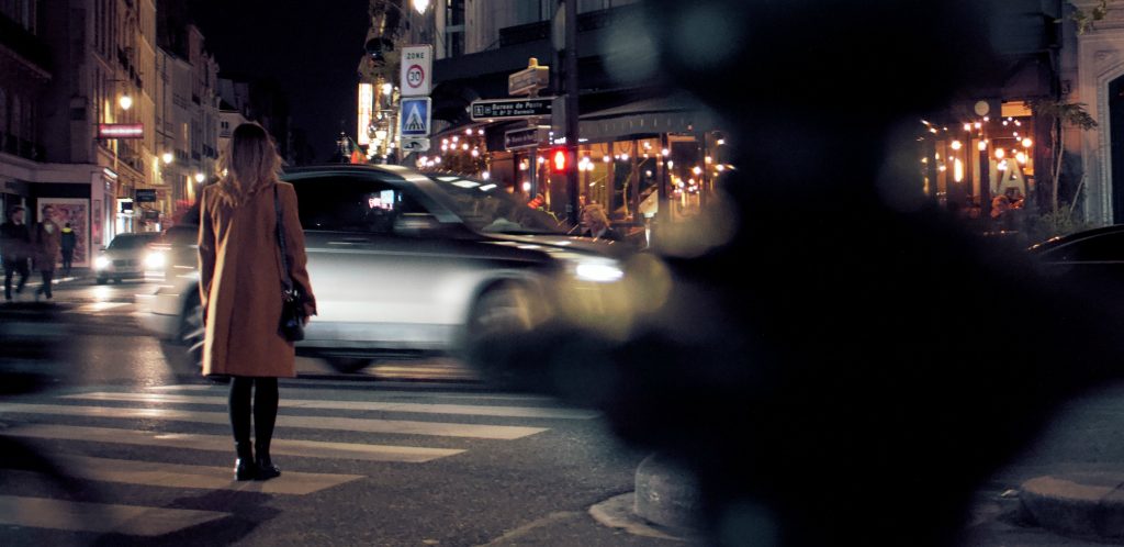 Woman in knee-length travel coat stands at crosswalk waiting for light to change in European city at night