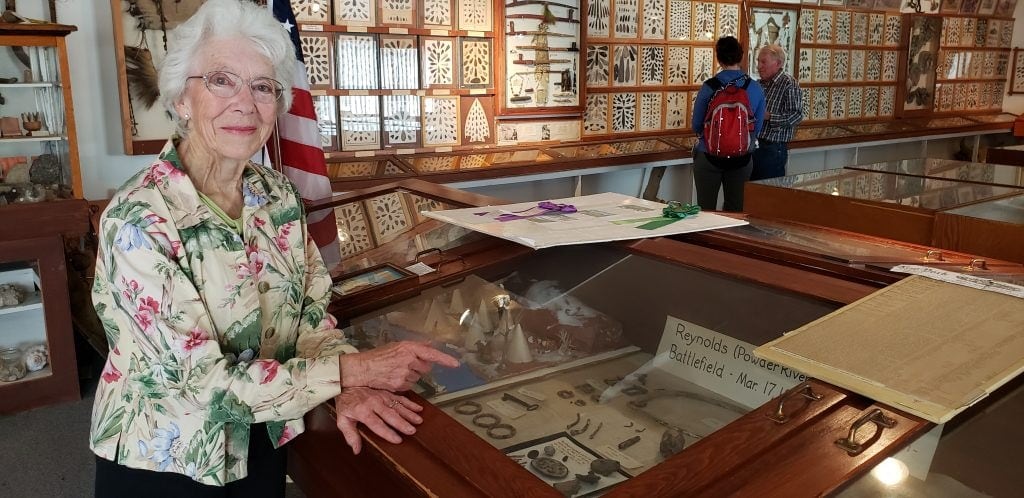 Silver-haired woman points to display case of authentic Wild West artifacts. Behind her displays line the walls.