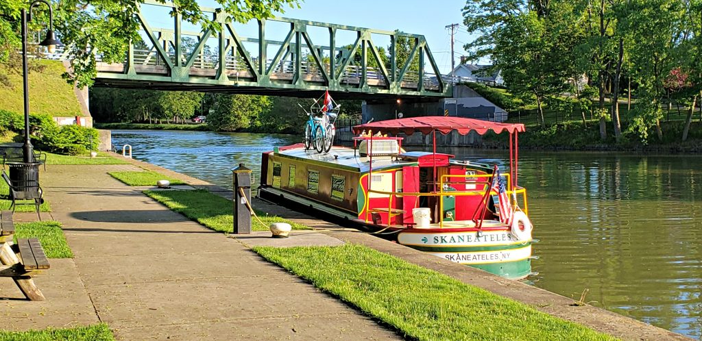 Red and green packet boat tied at pier on Erie Canal while a warren through truss bridge runs overhead.
