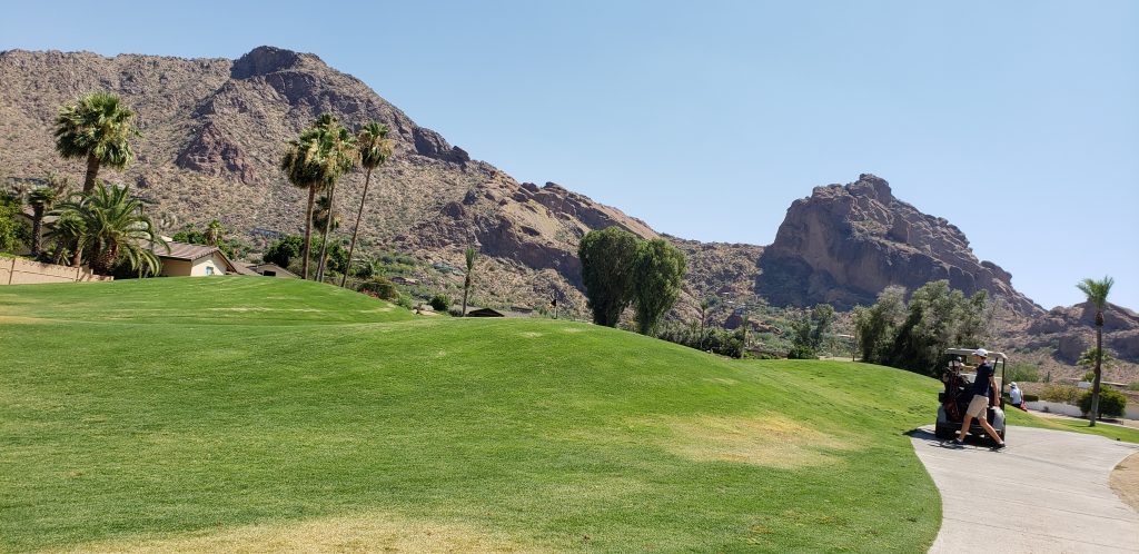 Golf cart on concrete path that weaves past a long green toward the reddish, pyramid-shape mountain in the background of the green golf course at Mountain Shadows golf course