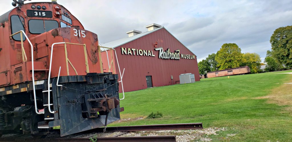 red locomotive in the foreground, train barn in background with sign reading "NAtional Trailroad Museum," which is One of the Best Green Bay WI Attractions