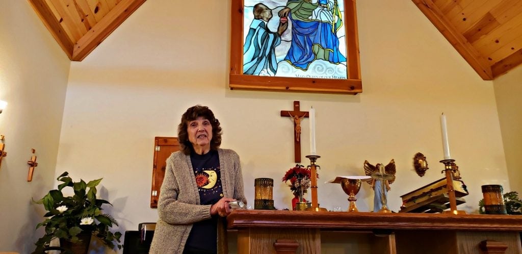 84 year old woman stands at altar of one of the Catholic shrines in Wisconsin - stained glass window above and cross on wall