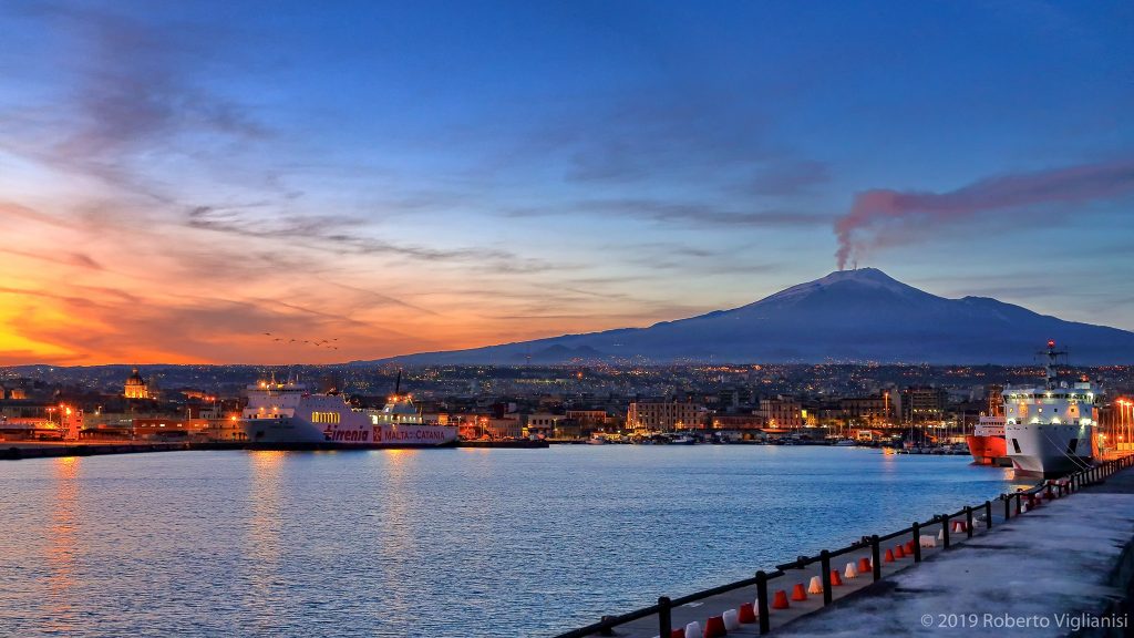 Harbor at sunset with silhouette of Mount Etna volcano steaming in the background