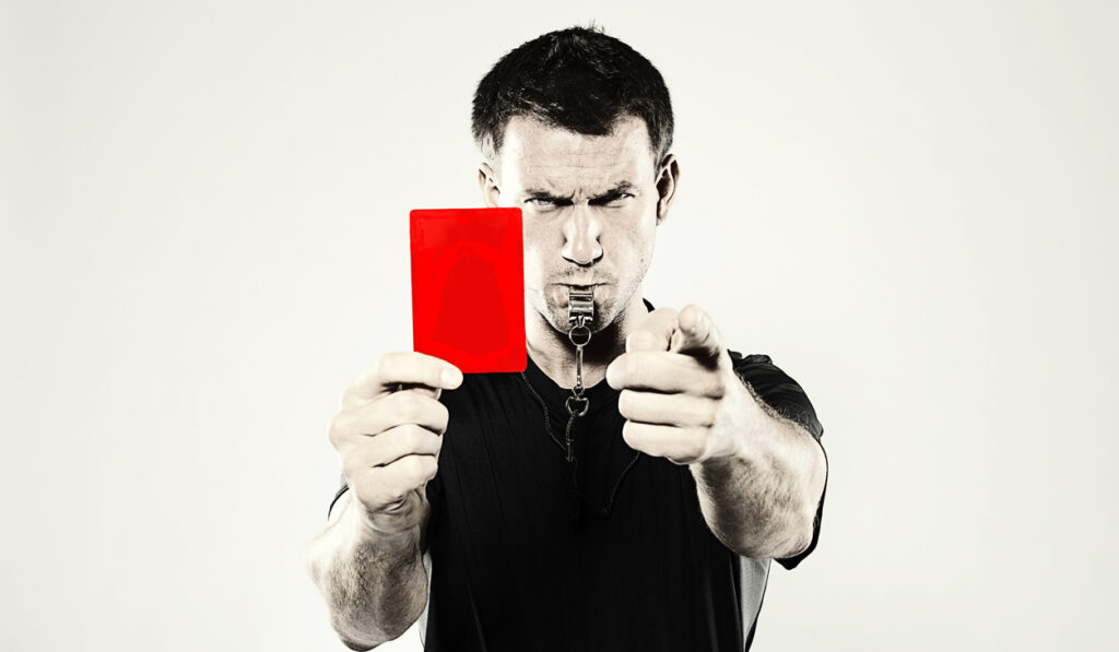 A male, Caucasian sports referee showing a red penalty card and blowing a whistle. Shot in the studio against a light background and with a retro photoshop effect.